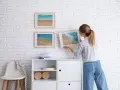 Mujer colocando cuadros de playa en pared de ladrillo blanco. Ideas para como combinar cuadros en la pared.