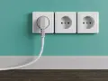 Tres enchufes blancos en una pared turquesa, uno con un enchufe gris conectado. La altura de los enchufes en la cocina es estándar.