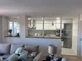 Un salón moderno con sofá y lámpara, separado de una cocina blanca y luminosa por una cristalera.