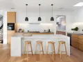 Cocinas modernas con isla blanca, taburetes de madera y gabinetes de roble. Escalera de diseño minimalista al fondo.