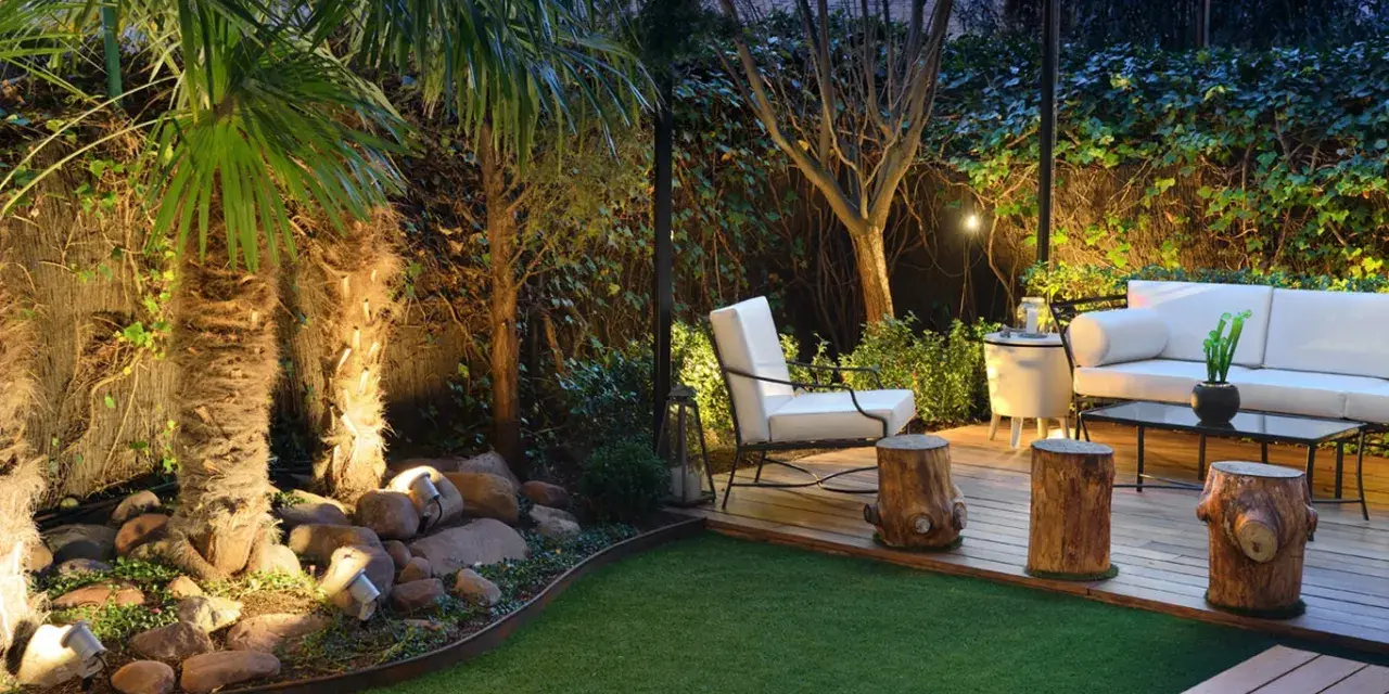 Un jardín poco mantenimiento con césped artificial, palmeras iluminadas y una zona lounge con sofá blanco y sillón.