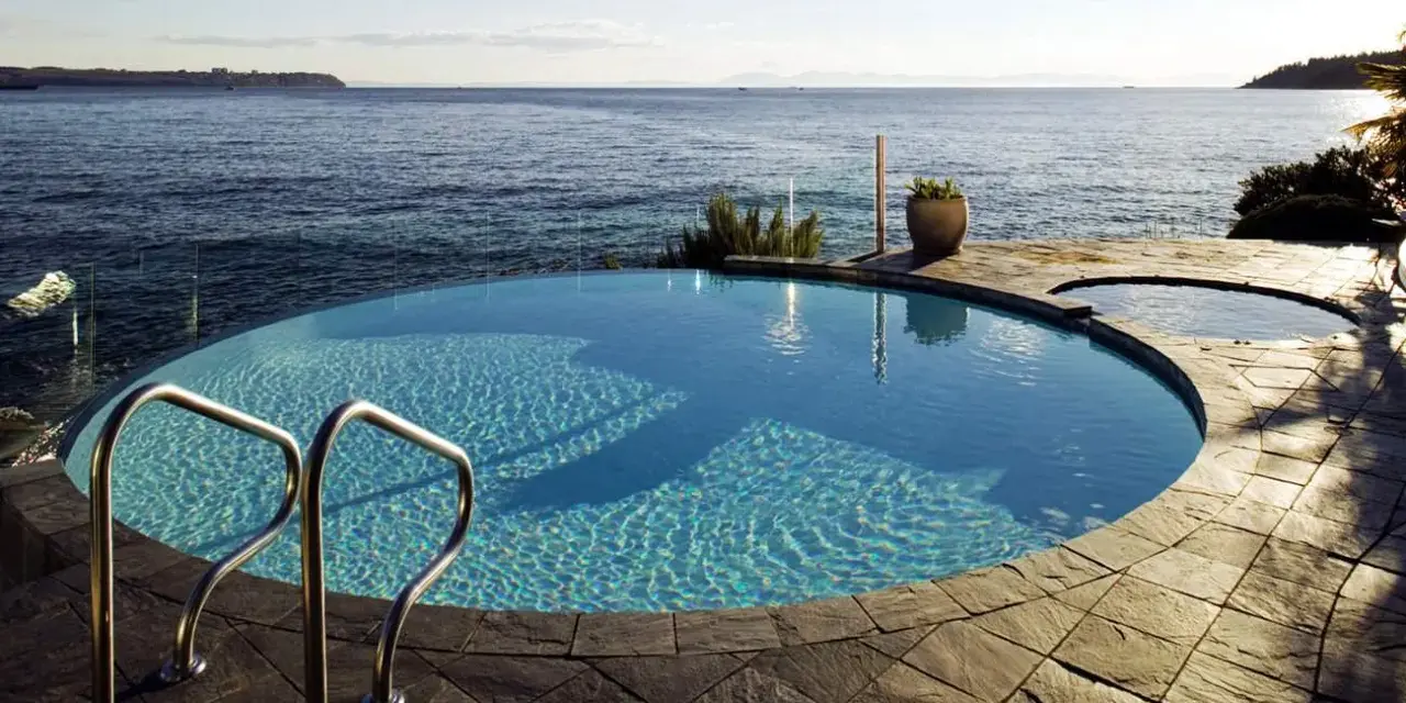 Una piscina infinita azul turquesa con escaleras de metal se funde con el océano azul bajo un cielo despejado.