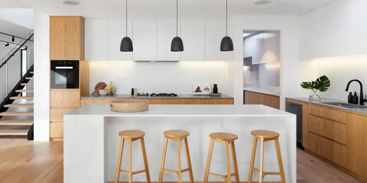 Moderna cocina blanca y madera con isla central, taburetes de madera y escaleras de diseño.