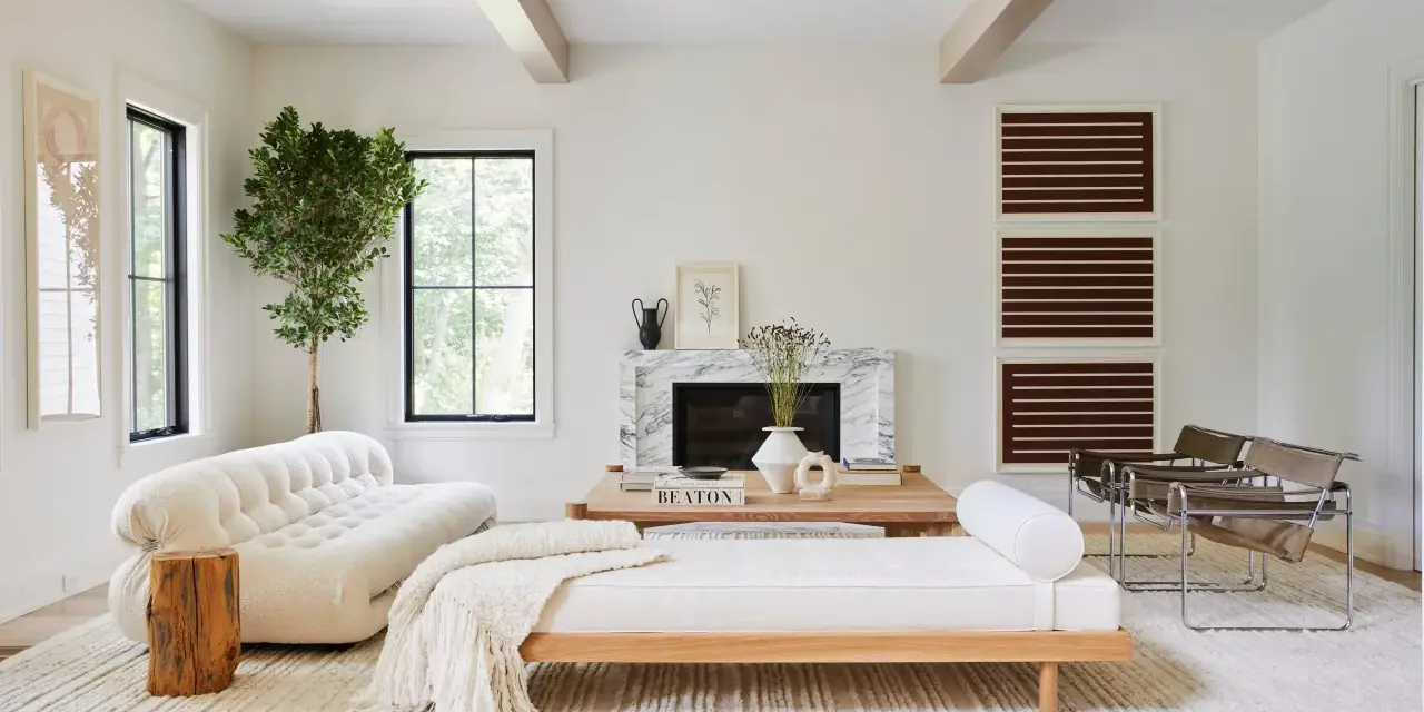 Un elegante salón blanco y madera natural, con sofá curvo, chaise longue, chimenea de mármol y sillas de cuero.