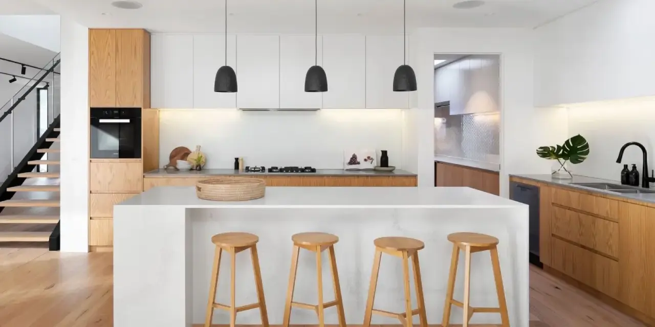 Cocinas modernas con isla blanca, taburetes de madera y gabinetes de roble. Escalera de diseño minimalista al fondo.