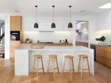 Cocinas modernas con isla blanca, taburetes de madera y gabinetes de roble. Escalera de diseño minimalista al fondo.