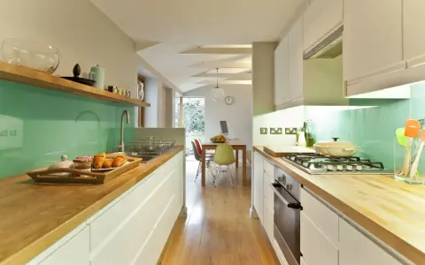 Cocina alargada con encimeras de madera, salpicadero verde turquesa y electrodomésticos integrados. Perfecta para distribuir el espacio.