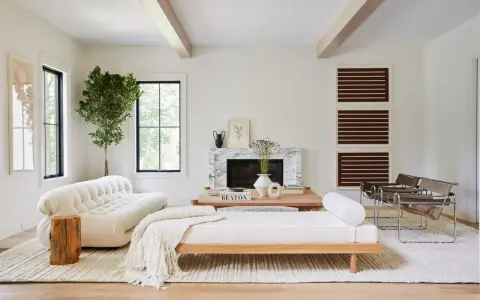 Un elegante salón blanco y madera natural, con sofá curvo, chaise longue, chimenea de mármol y sillas de cuero.