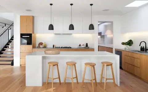 Cocinas modernas con isla blanca, taburetes de madera y gabinetes de roble. Escalera de diseño minimalista al fondo.