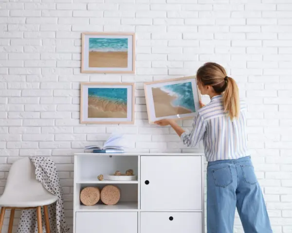 Mujer colocando cuadros de playa en pared de ladrillo blanco. Ideas para como combinar cuadros en la pared.