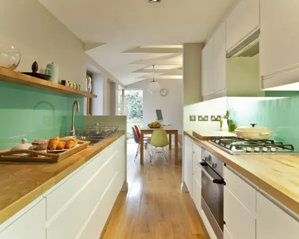 Cocina alargada con encimeras de madera, salpicadero verde turquesa y electrodomésticos integrados. Perfecta para distribuir el espacio.