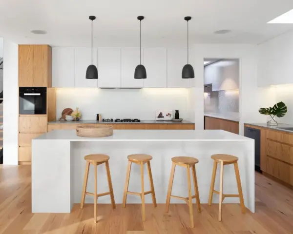 Cocinas modernas con isla blanca, taburetes de madera y gabinetes de roble. Escalera de diseño minimalista al fondo.