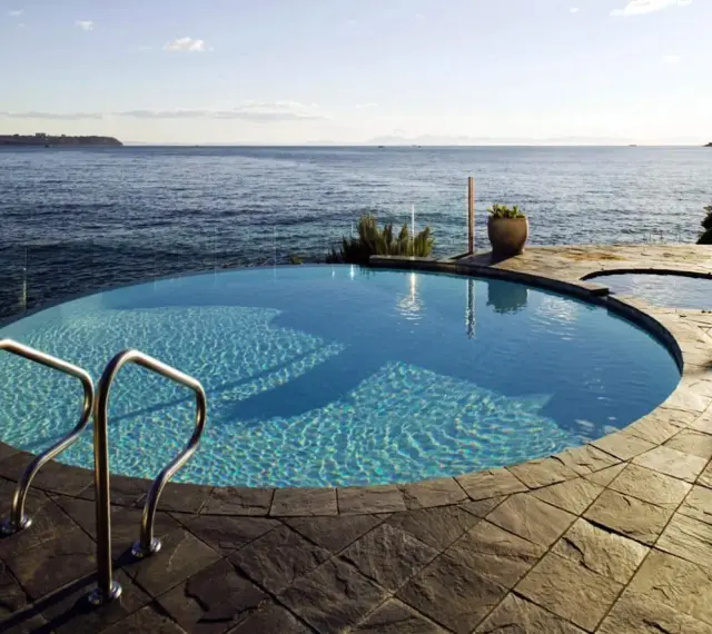 Una piscina infinita azul turquesa con escaleras de metal se funde con el océano azul bajo un cielo despejado.