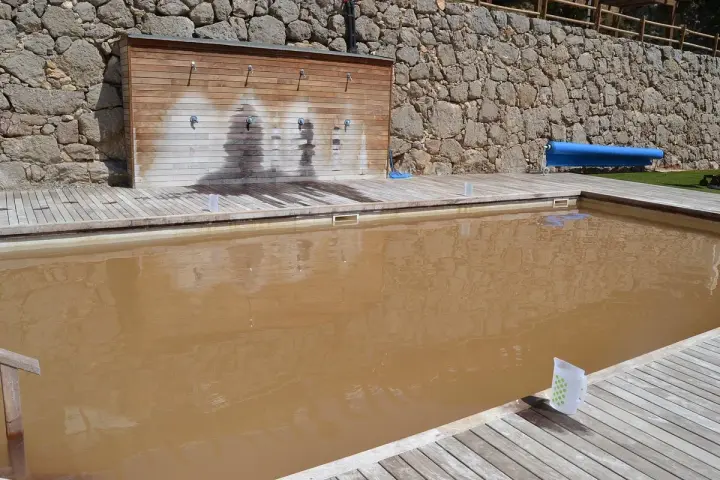 Piscina de sal con agua turbia y rodeada de madera. Al fondo, una pared de piedra y duchas.