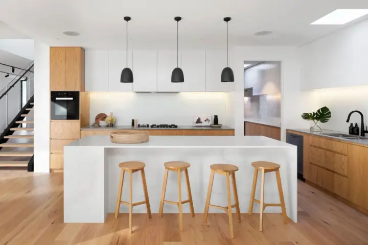 Cocinas modernas con isla blanca, taburetes de madera y gabinetes de roble. Escalera de diseño minimalista al fondo.