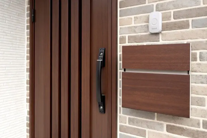 Puerta de madera oscura con tirador negro, buzón de madera y un timbre blanco. Varios tipos de timbre se pueden ver en la pared de ladrillos.