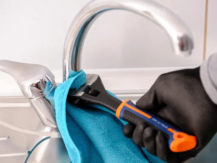 Mano enguantada usa llave inglesa para cambiar grifo cocina. Toalla azul protege el cromo.