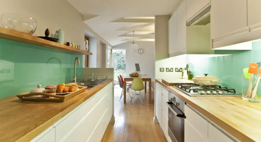Cocina alargada con encimeras de madera, salpicadero verde turquesa y electrodomésticos integrados. Perfecta para distribuir el espacio.