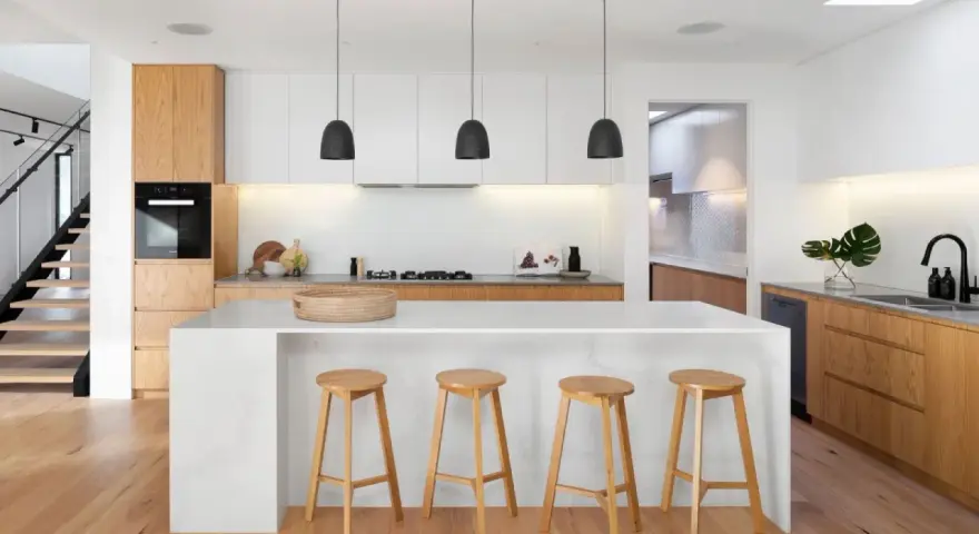 Cocinas modernas con isla blanca, taburetes de madera y gabinetes de roble. Escalera de diseño minimalista al fondo.