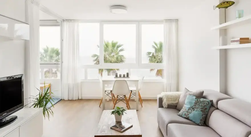 Acogedor salón nórdico rústico con sofá gris, mesa de centro de madera y vistas a palmeras.
