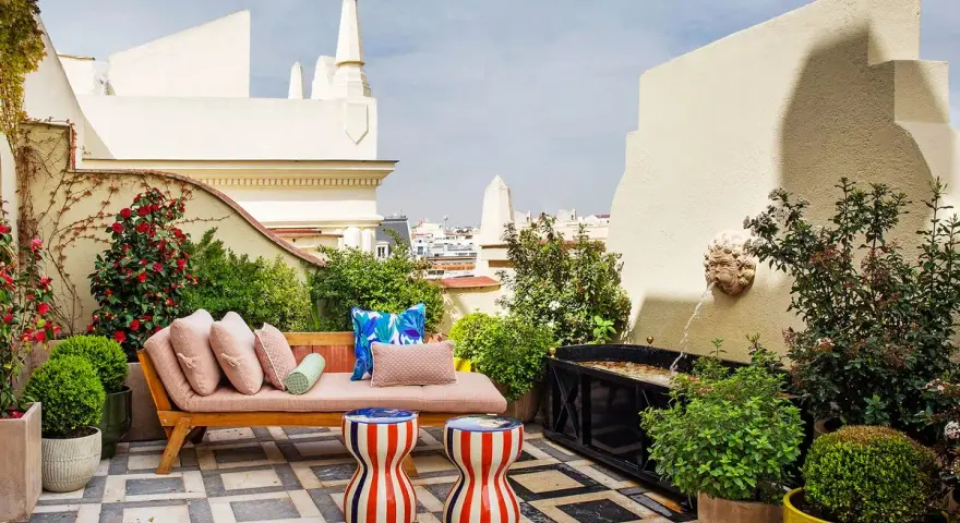 Sofá y taburetes coloridos en una terraza con fuente, ideal para decoracion terrazas pequeñas. Plantas y flores adornan este oasis urbano.