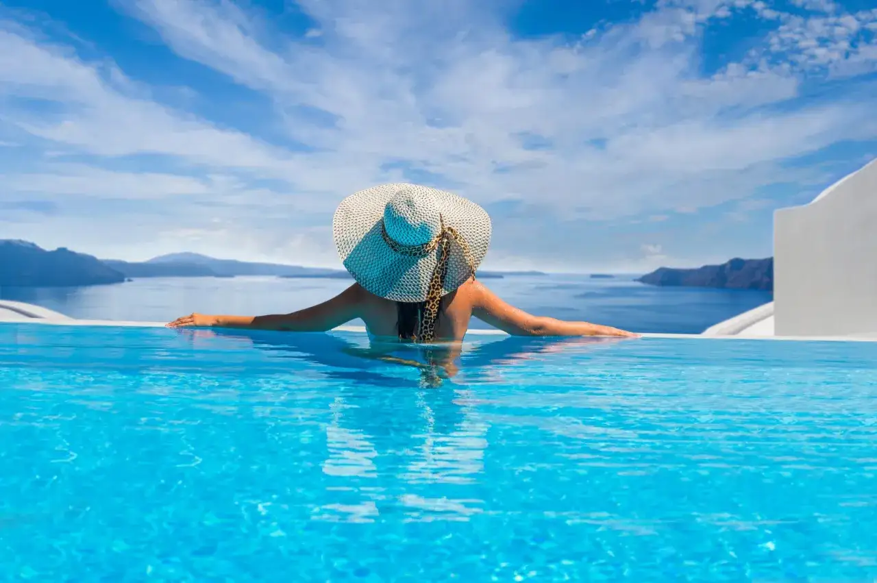 Mujer con sombrero disfruta de una piscina infinita con vistas al mar y montañas.