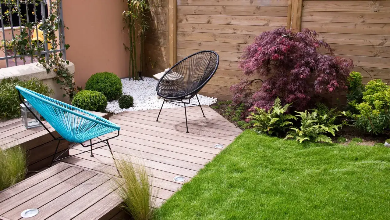 Dos sillas modernas en un patio con césped verde, plantas exuberantes y pequeños jardines cuidados.