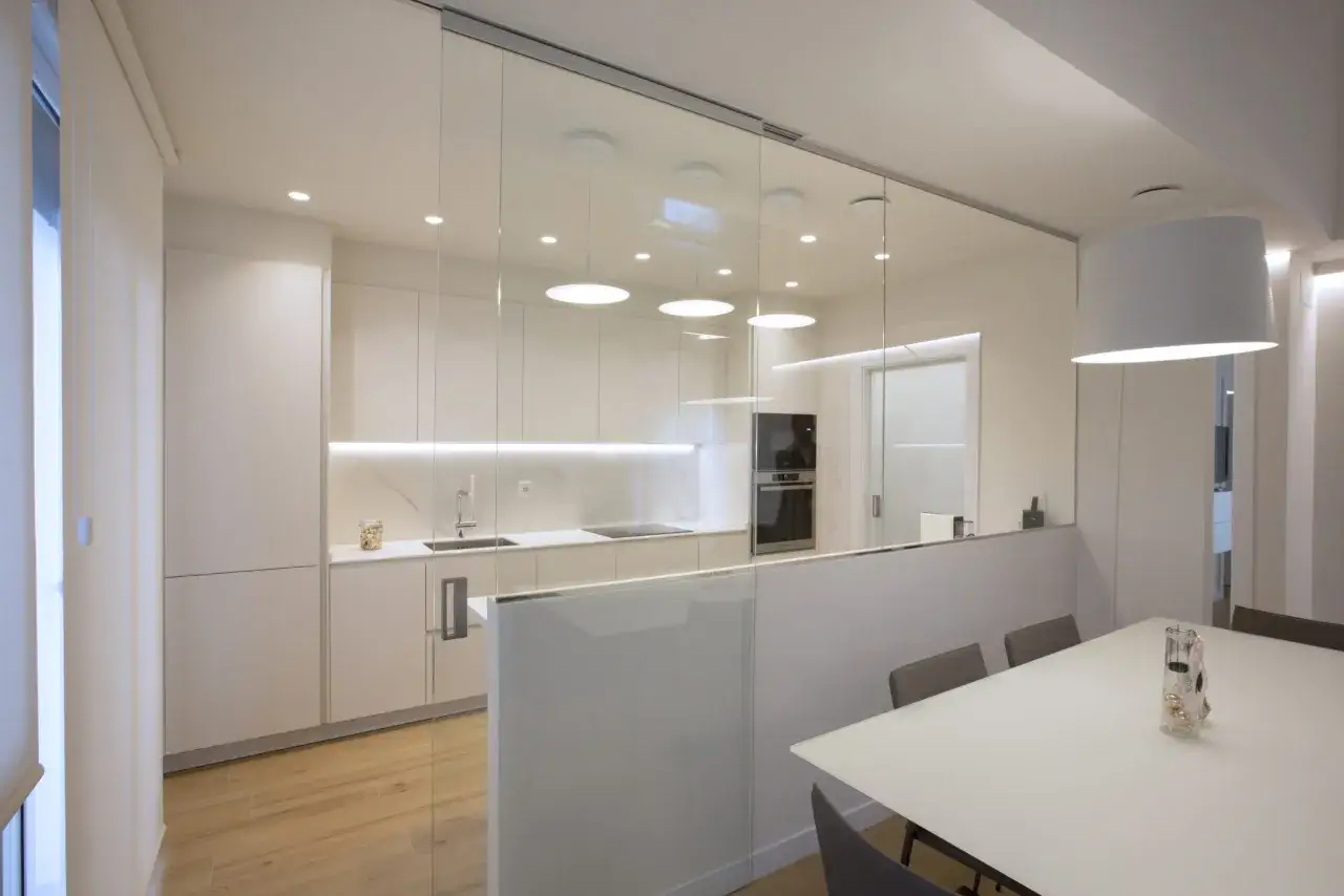 Cocina moderna con puertas de cristal correderas para separar cocina de salón. Muebles blancos, encimera de mármol y suelo de madera.
