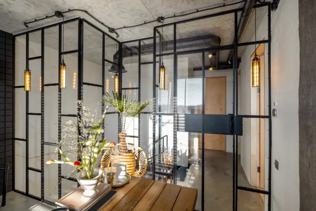Interior moderno con una pared de cristal que divide espacios. L&aacute;mparas colgantes y un arreglo floral sobre una mesa de madera.