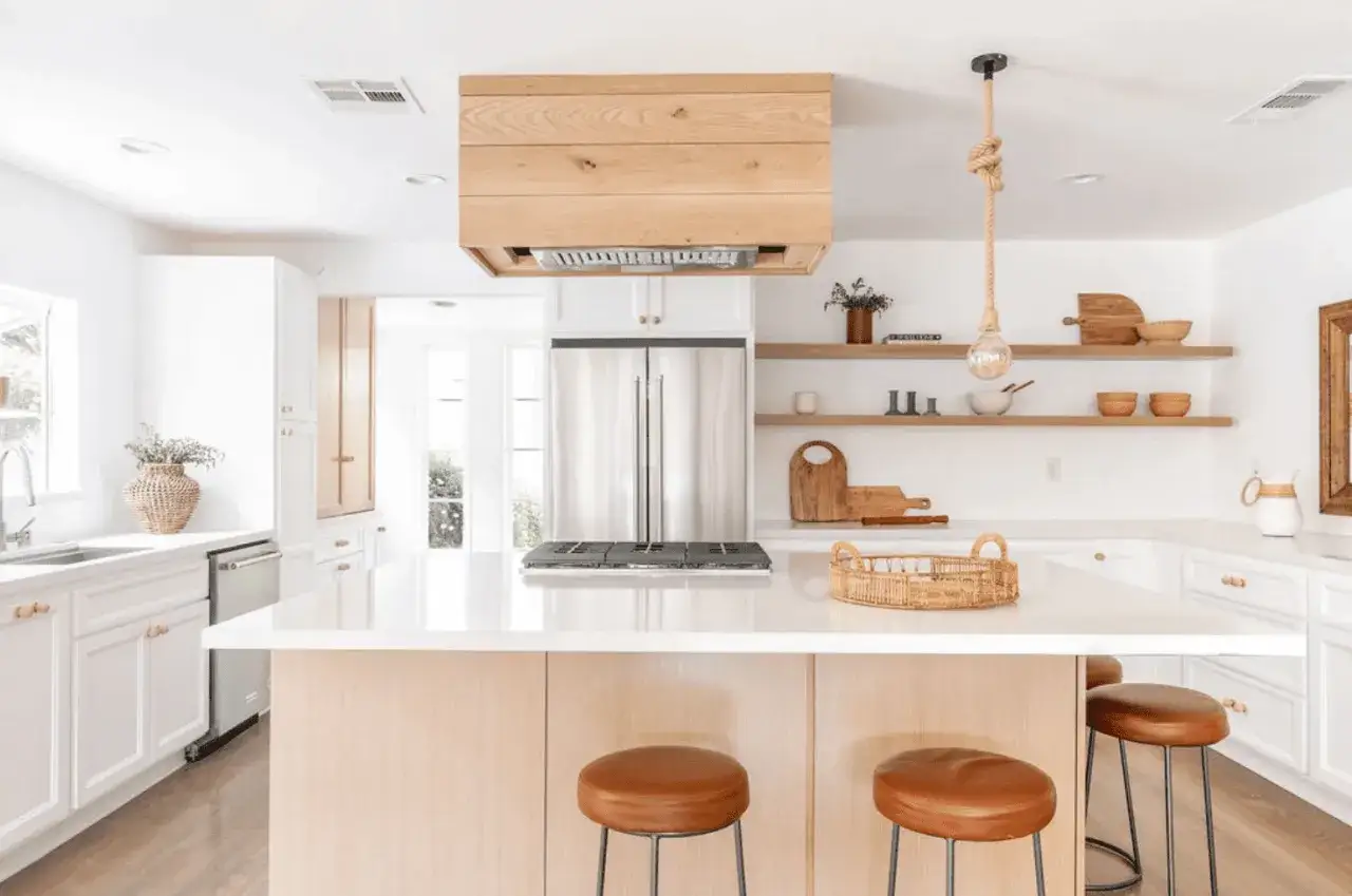 Cocinas color madera con isla central, encimeras blancas y taburetes de cuero. Estantes de madera con decoración rústica.