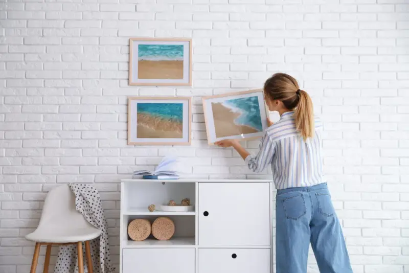 Mujer colocando cuadros de playa en pared de ladrillo blanco. Ideas para como combinar cuadros en la pared.