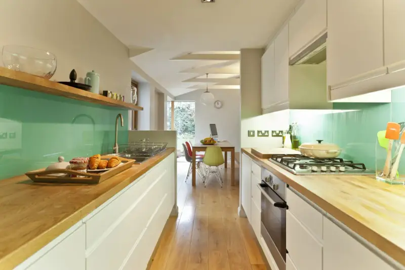 Cocina alargada con encimeras de madera, salpicadero verde turquesa y electrodomésticos integrados. Perfecta para distribuir el espacio.