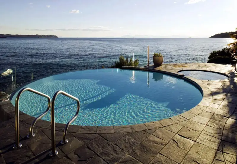 Una piscina infinita azul turquesa con escaleras de metal se funde con el océano azul bajo un cielo despejado.