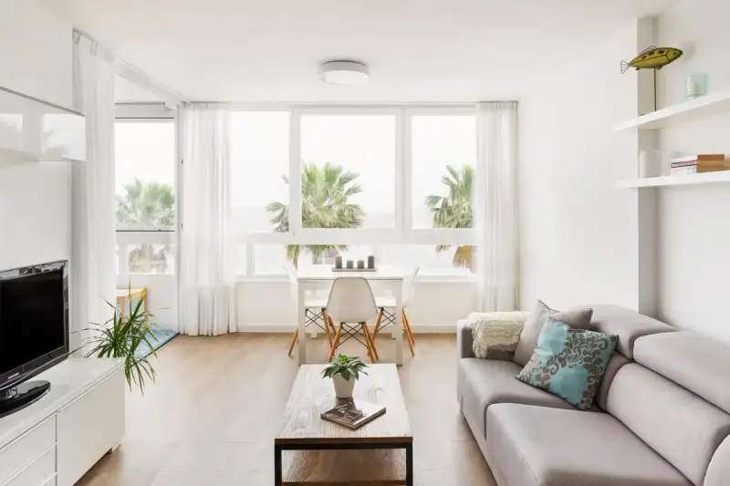 Acogedor salón nórdico rústico con sofá gris, mesa de centro de madera y vistas a palmeras.