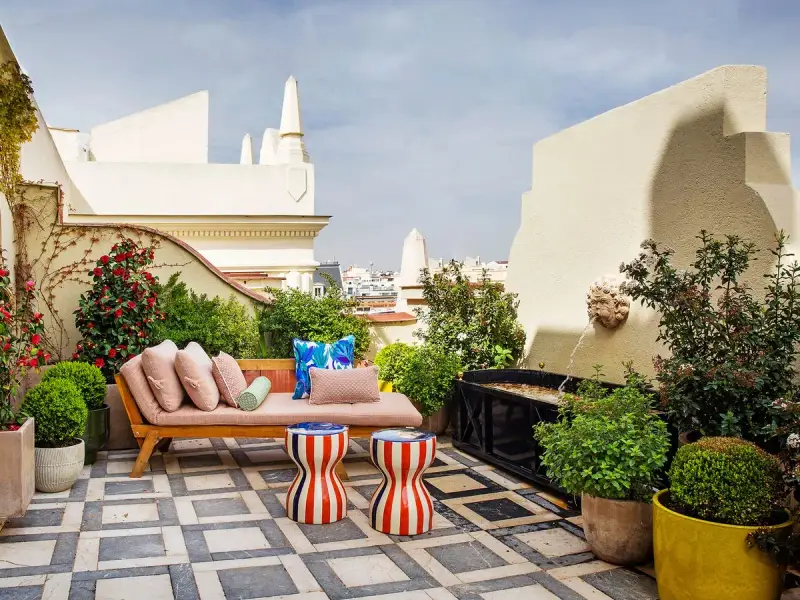 Sofá y taburetes coloridos en una terraza con fuente, ideal para decoracion terrazas pequeñas. Plantas y flores adornan este oasis urbano.